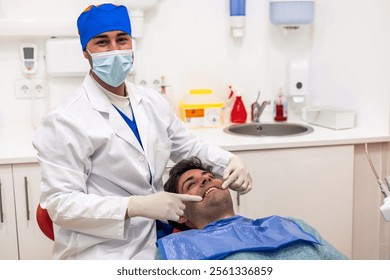 Young male dentist wearing a surgical mask, gloves, and a lab coat, pointing at a patient's teeth during a thorough dental checkup in a modern clinic setting - Powered by Shutterstock