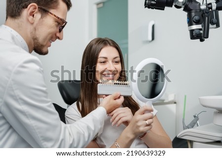 Young male dentist in white coat show veneers enamel color palette for beautiful patient woman sitting in a dental unit. Healthcare oral aesthetic treatment.