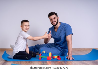 Young male doctor with beard in a blue uniform sitting on the floor next to the boy 10 years, they play educational toys, is looking at the camera and smile.