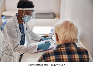 Young Male Doctor In A Face Shield And A Mask Examining Closely A Senior Man