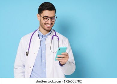 Young Male Doctor Looking At Smart Phone Screen With Smile, Exchanging Messages With Colleagues, Isolated On Blue Background, Copy Space On Right