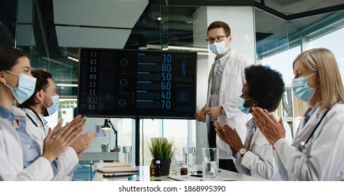 Young Male Doctor In Medical Mask Discuss With Mixed-races Healthcare Professional Coworkers Coronavirus Disease Statistics And Positive Results In Hospital While Colleagues Clapping Hands. Covid 19