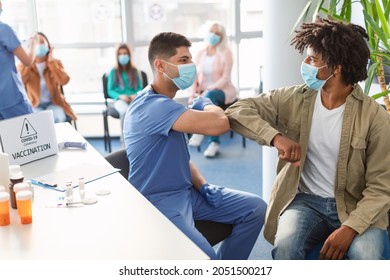 Young male doctor in medical protective face mask bumping elbows with African American man patient before vaccination against covid-19 virus during pandemic in medical clinic office vaccination point
