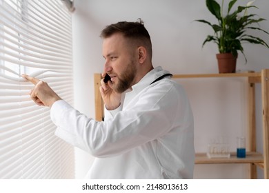 A Young Male Doctor Or Nurse Looks Out The Window, Parting The Blinds With His Fingers, Watching Something Outside And Talking On A Mobile Phone. Copy Space.