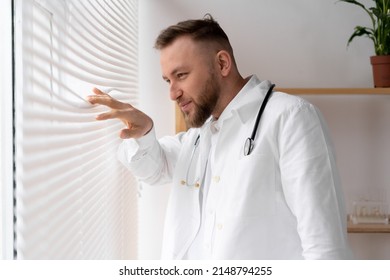 A Young Male Doctor Or Nurse Looks Out The Window, Parting The Blinds With His Fingers, Watching Something Outside. Copy Space.