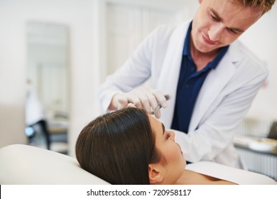 Young Male Doctor Performing A Botox Injection To The Forehead Of A Woman Lying On A Table In A Beauty Clinic