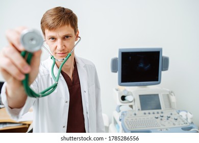 Young Male Doctor Pointing Stethoscope Head At Camera, Looking At You. He's Standing Next To Ultrasound Scanning Machine.