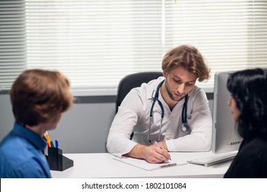 A Young Male Doctor Receiving A Teen Patient With His Parent At The Office, He Is Asking Questions And Taking Notes.