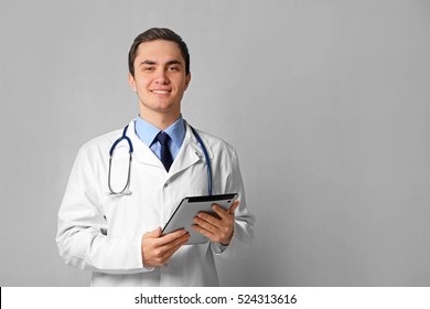 Young male doctor with tablet on grey wall background