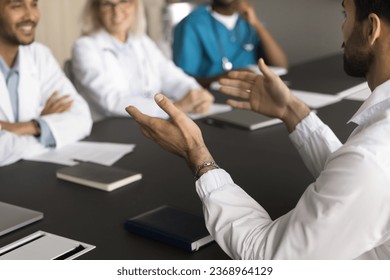 Young male doctor talking to diverse team of cheerful positive colleagues. Medical practitioners meeting for collaboration, discussing successful job cases, healthcare issues at conference table - Powered by Shutterstock