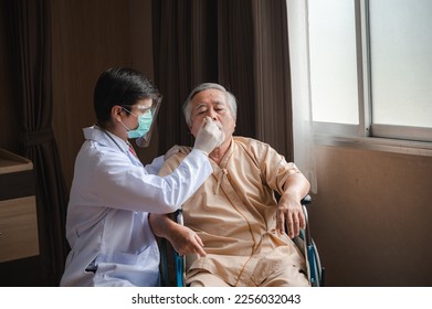 Young male doctor wearing lab coat with face shield mask and surgical mask for safety against covid-19 measuring temperature of senior old man sitting on wheelchair using thermometer in hospital room - Powered by Shutterstock