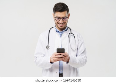 Young Male Doctor In White Coat Smiling While Looking At Screen Of His Phone, Using Medical App, Standing Isolated On Gray Background
