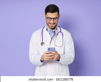 Young Male Doctor In White Coat Smiling While Looking At Screen Of His Phone, Using Medical App, Standing Isolated On Purple Background