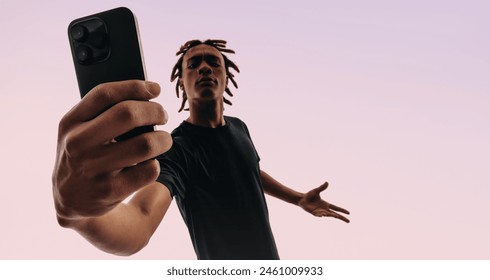 Young male with dreadlocks holds a pink smartphone, taking a self portrait with a selfie camera. Gen Z in a studio background, showcasing their use of mobile apps for social media. - Powered by Shutterstock