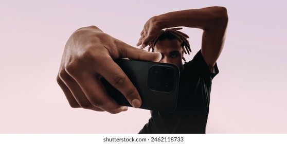 Young male with dreadlocks takes a selfie using a cellphone app. He strikes a pose in a pink studio, showcasing his Gen Z lifestyle and love for technology and social media. - Powered by Shutterstock