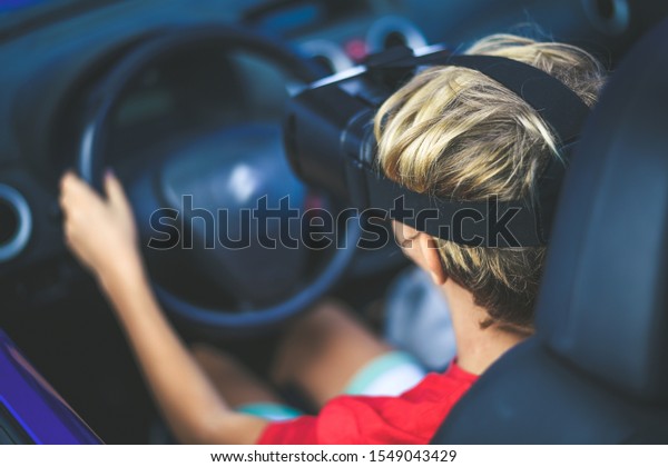 Young male drive a car with virtual reality\
goggles. Boy sitting behind the steering wheel simulates driving a\
car with new tech devices. Learning road safety with vr headset.\
New technology concept