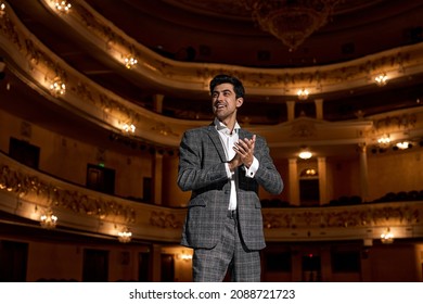 Young Male Entertainer, Presenter Or Actor On Stage. Male Public Speaker Speaking At Microphone, Performing Presentation On Stage, Looking At Audience. Guy In Classic Formal Wear Clapping Hands