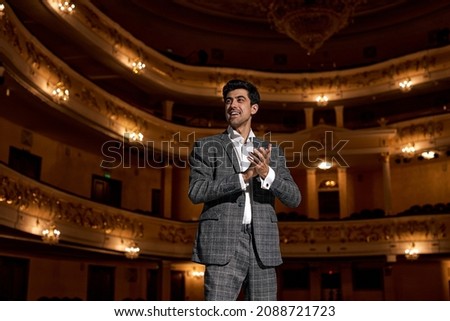 Young male entertainer, presenter or actor on stage. male public speaker speaking at microphone, performing presentation on stage, looking at audience. guy in classic formal wear clapping hands