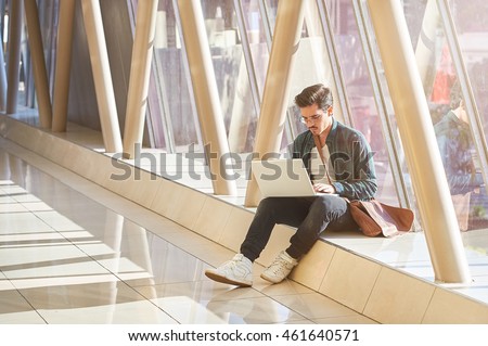young male entrepeneur student waiting working on laptop in sunny corridor