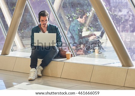 young male entrepeneur student waiting working on laptop in sunny corridor