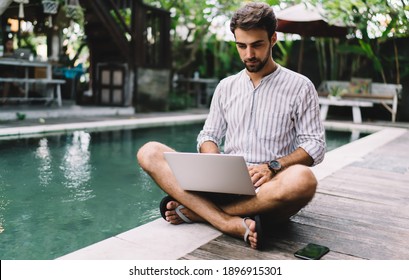 Young male entrepreneur in summer shirt and flip flops sitting on poolside and using laptop to do online business project in quiet yard of resort house