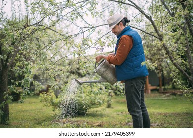 A Young Male Farmer In Work Clothes Is Watering Plants From A Watering Can In His Garden. A Guy In A Work Shirt And Vest With A Metal Fishing Line In His Hands