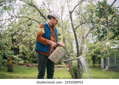 A Young Male Farmer In Work Clothes Is Watering Plants From A Watering Can In His Garden. A Guy In A Work Shirt And Vest With A Metal Fishing Line In His Hands