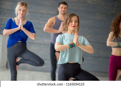 Young Male And Female Students Doing Yoga Exercise In College Sport Hall Standing In Tree Pose, Vrksasana Posture, Taking Off Tension Of Exams Time