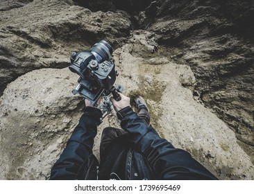 Young male filmmaker photographer pov hold dslr camera in hands and using stabilizer for pictures in mountains with beautiful landscape standing in sand in snickers and black coat