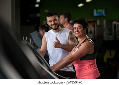 Young Male Fitness Coach With Middle Aged Female Client On Treadmill Machine. Attractive Brunette Woman Working Out In Gym With Personal Trainer. Healthy Lifestyle Concept. Selective Focus