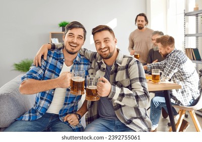 Young male friends having fun and drinking beer at party at home. Portrait of two happy men in plaid shirts sitting on couch at home, hugging, holding beer mugs, looking at camera, and smiling - Powered by Shutterstock