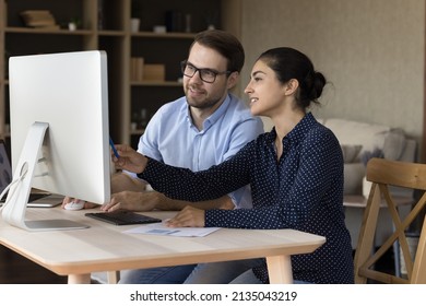 Young Male In Glasses Trainee Apprentice Take Professional Internship Work By Desktop Computer In Team With Experienced Indian Female Mentor. Two Colleagues Of Diverse Gender Race Interacting Using Pc