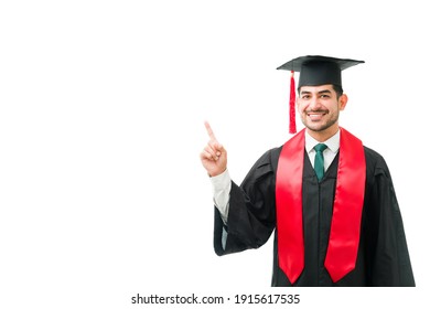 Young Male Graduate Wearing A Black Gown, Cap And Red Tassel Against A White Background. Attractive Man Smiling And Pointing To A Copyspace