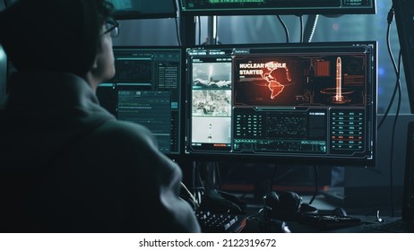 Young Male Hacker In Hat And Glasses Using Computers To Hack Nuclear Warhead And Start War While Sitting At Table In Dark Room