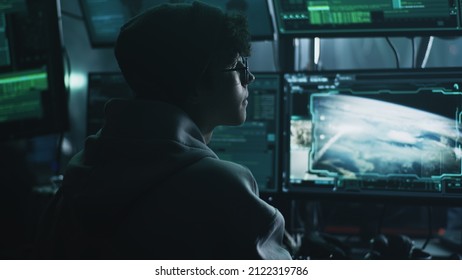 Young Male Hacker In Hat And Glasses Using Computers To Hack Nuclear Warhead And Start War While Sitting At Table In Dark Room