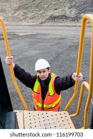 A Young Male Heavy Equipment Operator Climbing Up The Steps Of A Loader Machine At A Construction Site.