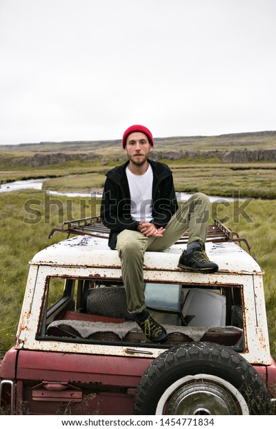 Young\
male hipster or social media blogger poses on top of remains of old\
rotten car at field or old vehicle junk yard. Looks at camera.\
Explore unknown and look cool in strange\
scenery