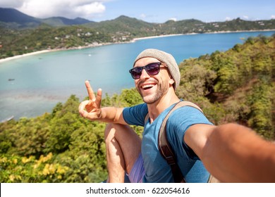 Young male hipster traveler doing selfie overlooking the tropical sea. Adventure, vacation, wonderlust, internet, technology concept.