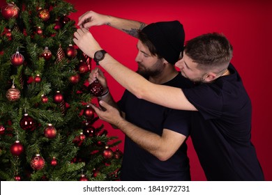 Young Male Homosexual Couple Decorating Xmas Tree Together Celebrating New Year, Two Stylish Gay Men Preparing For Family Party. Merry Christmas, Winter Holiday Concept. Red Background