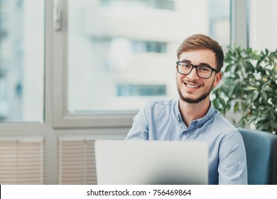 Young male intern with glasses working on laptop and smiling at the camera