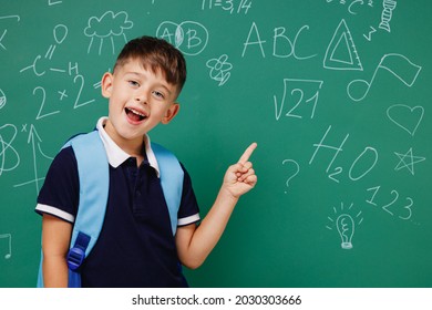 Young Male Kid School Boy 5-6 Years Old In T-shirt Backpack Point Finger Aside On Workspace Area Isolated On Green Wall Chalk Blackboard Background. Childhood Children Kids Education Lifestyle Concept