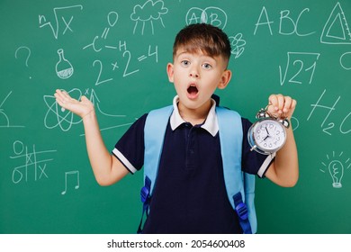 Young Male Kid School Boy 5-6 Years Old Wear T-shirt Backpack Hold In Hands Clock Spread Hand Isolated On Green Wall Chalk Blackboard Background. Childhood Children Kids Education Lifestyle Concept