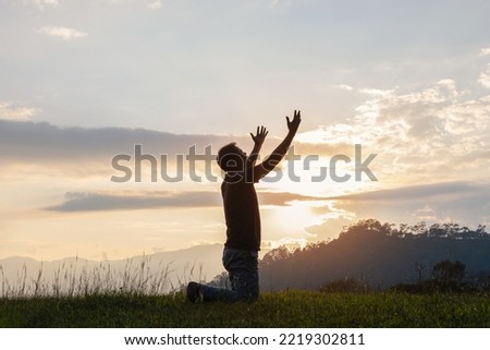 Young male kneeling down with hands open palm up praying to God on the mountain sunset background. 