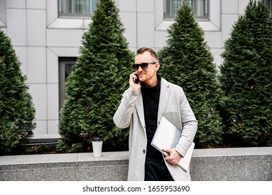 Young Male Landscape Designer With Laptop Waiting For A Meeting With A Client. He Is Talking On A Smartphone.