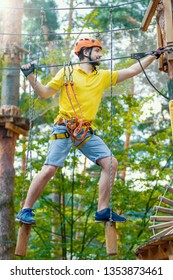 Young Male Man Adult Wears Protective Helmet With Action Camera Having Fun In Extreme Rope Park, Amusement Park. Climbing In Rope Bridge At Green Forest. Active Healthy Lifestyle In Spring Or Summer.