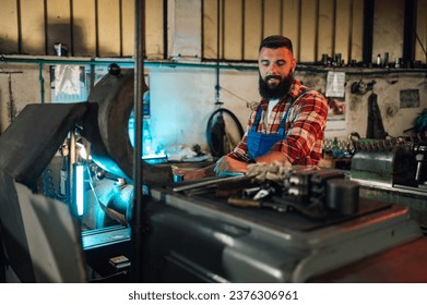 Young male metallurgist with a beard working on a milling machine to create a new metal part. He wears a blue worker uniform and a plaid shirt. Old workshop, manufacture, artisan, craftsman. - Powered by Shutterstock