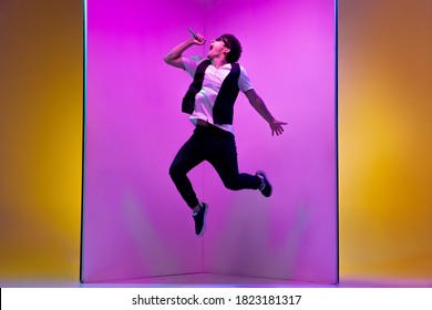 Young Male Musician, Singer Performing On Pink-orange Background In Neon Light. Concept Of Music, Hobby, Festival, Entertainment, Emotions. Joyful Party Host, Singer, Portrait Of Artist.