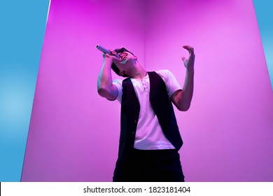 Young male musician, singer performing on pink-blue background in neon light. Concept of music, hobby, festival, entertainment, emotions. Joyful party host, singer, portrait of artist. - Powered by Shutterstock