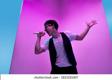 Young Male Musician, Singer Performing On Pink-blue Background In Neon Light. Concept Of Music, Hobby, Festival, Entertainment, Emotions. Joyful Party Host, Singer, Portrait Of Artist.