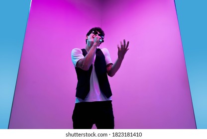 Young Male Musician, Singer Performing On Pink-blue Background In Neon Light. Concept Of Music, Hobby, Festival, Entertainment, Emotions. Joyful Party Host, Singer, Portrait Of Artist.